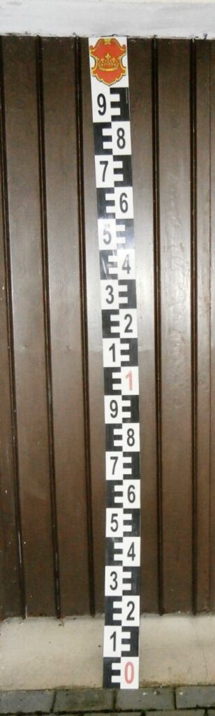 2,2 metru dlouhá hladinová vodočetná lať typ III. s logem obce pro AQUATEST opřená o zeď, připravena na expedici